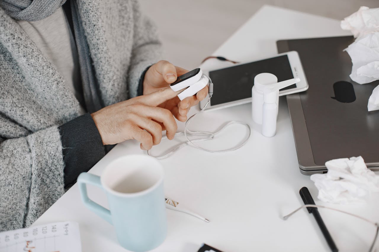 Adult using a pulse oximeter at a home desk, surrounded by tech and healthcare items.